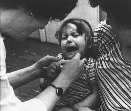 Varšuvos vaikų sveikatos klinikos slaugytoja bando sušvirkšti jodo tirpalą trejų metų mergaitei, laikomai motinos rankose. 1986 m. gegužės mėn. Varšuvos vaikų sveikatos klinikos slaugytoja bando sušvirkšti jodo tirpalą trejų metų mergaitei, laikomai motinos rankose. 1986 m. gegužės mėn.