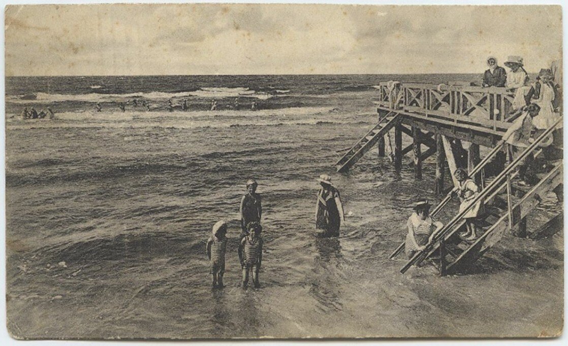 Moterys ir vaikai maudosi prie Palangos jūros tilto, 1905 m. Damos dėvi maudymosi sukneles ir skrybėles, o vaikai – uždarus maudymosi kostiumus. Paulinos Mongirdaitės atvirukas.